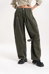 Pantalón cargo de tiro medio-alto, color verde militar, con diseño holgado. Presenta múltiples bolsillos laterales, cierres decorativos y funcionales en la parte frontal y una tira ajustable en una de las piernas.