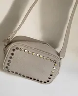 Cartera bandolera de cuero sintético color beige, con diseño rectangular y detalle de tachas metálicas circulares en el contorno frontal. Cuenta con correa larga ajustable y desmontable mediante mosquetones, cierre superior y bolsillos internos y externos.