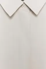 Camisa de corte fluido con cuello solapa y manga corta con dobladillo. Presenta cierre frontal con botones ocultos bajo una tapeta.