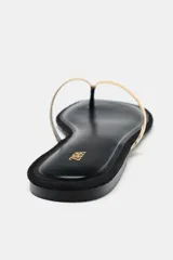 Sandalia plana tipo ojota, color negro, con tira entre los dedos y una tira superior ancha y curva de acabado metálico dorado.