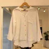 Camisa de lino blanca de manga larga con cuello camisero, cierre frontal con botones y un bolsillo aplicado en el pecho. Presenta diseño de corte asimétrico con dobladillo redondeado más largo en la parte trasera.