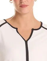 Blusa de crepe negro con ribetes en color beige que recorren el escote, la línea central y las costuras de los hombros. Presenta cuello redondo con abertura en V, mangas largas con puños abiertos y detalle de botón metálico, y ruedo redondeado.