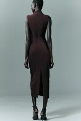 Vestido midi de color negro, con cuello subido y diseño sin mangas. Presenta un detalle de drapeado lateral en la parte delantera y cierre de cremallera oculta en la espalda.