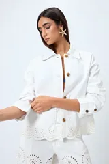 Sobrecamisa blanca de corte recto con cuello clásico, cierre frontal de botones y bolsillos delanteros. Presenta un delicado detalle de bordado inglés en el ruedo.