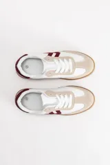 Zapatilla de estilo urbano con diseño acordonado, confeccionada en una combinación de cuero sintético blanco, detalles en gamuza beige y apliques laterales en color bordó. Cuenta con suela de goma tipo caramelo.