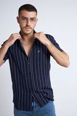 Camisa de manga corta con diseño a rayas verticales en color azul marino y blanco. Presenta cuello clásico, cartera abotonada frontal y logo bordado en el pecho. Posee un corte slim fit.