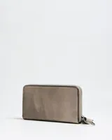 Billetera grande tipo fichero de cuero sintético color taupe, con cierre perimetral, detalles de tachas metálicas plateadas en los bordes y placa frontal con la inscripción de la marca Extra Large. Incluye una tira de mano desmontable con argolla metálica.