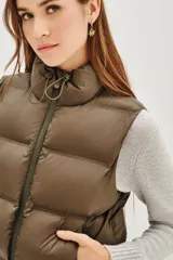 Chaleco acolchado de corte corto, color verde oliva, con cuello alto ajustable mediante cordones elásticos y cierre frontal con cremallera.