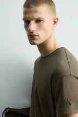 Camiseta de algodón color verde oliva, corte regular fit, con cuello redondo y manga corta. Presenta un pequeño corazón bordado en la manga.