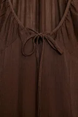 Vestido midi de color marrón, confeccionado en tejido de gasa con textura. Presenta un escote en pico con detalle de lazada frontal, mangas cortas abullonadas con terminación elástica y falda con detalle de godets que aportan volumen. Incluye forro interior a tono y cierre lateral con cremallera oculta.