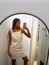 Vestido corto de lino en color beige, con escote cuadrado y breteles anchos. Presenta pespuntes decorativos y tajos laterales para mayor comodidad y movimiento.