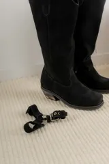 Bota estilo tejana de caña alta, confeccionada en cuero marrón con acabado texturizado. Presenta punta cuadrada, taco bajo y una correa con hebilla metálica ajustable y estribo desmontable en la zona del tobillo.