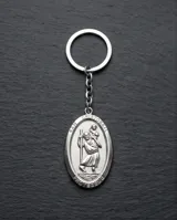 Llavero de acero con dije ovalado que presenta un relieve de San Cristóbal, santo patrono de los viajeros, con argolla circular de doble vuelta.