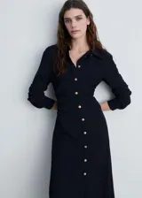 Vestido camisero de color negro con diseño entallado y falda evasé. Presenta cuello camisero, cierre frontal con botones dorados, mangas largas con puños abotonados y detalle de cintura drapeada.