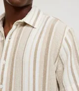 Camisa de hombre de manga corta con cuello tradicional y abotonado frontal. Presenta un tejido texturizado con un patrón de rayas verticales en tonos beige, blanco y marrón claro.