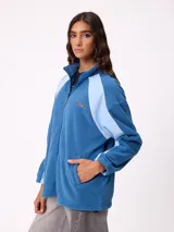 Campera de polar marca Rusty, color azul con paneles laterales en celeste claro. Presenta cierre frontal completo, cuello alto y logo de la marca bordado en el pecho.