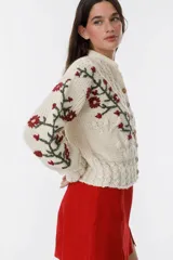 Cárdigan tejido a mano color crudo, con diseño de trenzas y flores bordadas en azul y marrón. Tiene cuello redondo y cierre frontal con botones de madera.