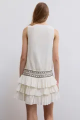 Vestido corto de gasa color blanco, con escote en V cruzado, sin mangas y diseño con volantes en el bajo. Presenta un detalle de tachas plateadas en la cintura.