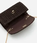 Cartera pequeña de rafia color marrón oscuro con textura de efecto paja. Posee una correa de cadena dorada para uso transversal y un cierre frontal de metal tipo locker.
