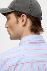 Camisa de corte relajado confeccionada en algodón, con diseño de rayas verticales en tonos azul, blanco y rojo. Presenta cuello americano, cierre frontal con botones, bolsillo en el pecho y mangas largas con puños abotonados.