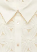 Blusa color crema con textura calada de flores en relieve, cuello camisero, manga corta y cierre de botones en el cuello.