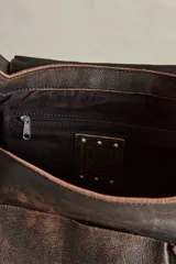 Cartera hobo de cuero color marrón oscuro con acabado gastado. Presenta una solapa frontal con detalles de ojales metálicos, una argolla metálica en la correa para el hombro y bolsillos laterales. Incluye un llavero colgante con candado y dijes.