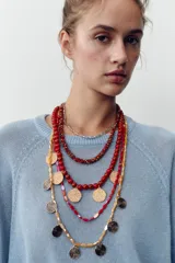 Pack de cinco collares superpuestos con cadenas de diferentes grosores. Incluye collares con cuentas rojas, marrones y amarillas, cuentas de cristal transparentes/rojas, y dijes metálicos circulares con textura martillada dorada y otros con diseño de espiral plateado/negro.