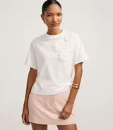 Remera blanca de algodón con cuello redondo y corte boxy, con apliques de flores de tejido en relieve en la zona del pecho.