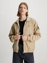 Campera bomber color beige de nailon reciclado, con cuello alto de canalé, puños y bajo elásticos, cierre de cremallera, bolsillos con botón de presión en la cintura, forro de poliéster reciclado e insignia con monograma de Calvin Klein en la manga.