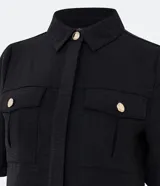 Camisa negra de manga corta con cuello inglés, dos bolsillos delanteros con solapa y botones dorados. Presenta un tejido texturizado y un dobladillo trasero más largo (barra mullet).