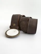 Cinto de cuero color chocolate oscuro con forro de cuero. Presenta una hebilla circular con borde metálico labrado y centro de guampa, con sistema de broche suizo intercambiable.