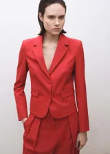 Chaqueta tipo blazer de corte recto y diseño estructurado, color rojo intenso. Presenta cuello con solapas de muesca, cierre frontal de dos botones, mangas largas con puños abotonados y dos bolsillos laterales con solapa.