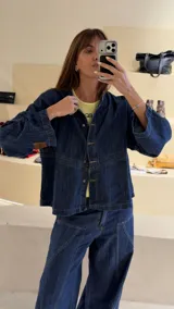 Camisa de denim azul con cuello mao, diseño holgado, mangas largas y cierre frontal con botones metálicos. Cuenta con costuras decorativas pespunteadas en el delantero.