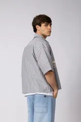 Camisa de manga corta de corte boxy, confeccionada en popelina a rayas celestes y blancas. Presenta cuello camisero clásico, cierre frontal con botones y estampado tipográfico en el pecho.
