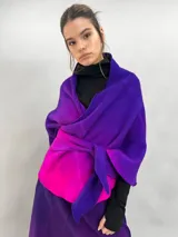 Prenda de abrigo tipo capa o chal de lana merino afieltrada, con diseño desestructurado y efecto degradé en tonos violeta y fucsia. Se ajusta al cuerpo mediante un nudo frontal.