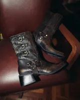 Bota de cuero vacuno color marrón oscuro, de caña media, con punta cuadrada y taco bajo. Presenta correas con hebillas metálicas plateadas y estribo desmontable.