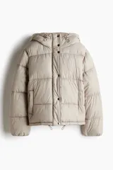 Campera puffer color beige con capucha, cierre frontal y solapa con botones a presión. Cuenta con bolsillos laterales, elástico en los puños y cordón ajustable en la capucha y el ruedo.