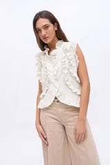 Blusa sin mangas de lino color beige, con cuello alto y detalles de volados en los hombros y a lo largo de la botonera frontal.