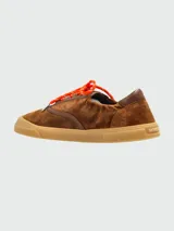 Zapatillas Moncler modelo Vera Leisure, confeccionadas en gamuza marrón con detalles en cuero tono sobre tono. Presentan cordones en color naranja vibrante y suela de goma texturizada en color caramelo.
