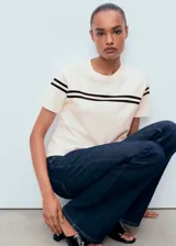 Jersey de punto fino color crudo, con cuello redondo y mangas cortas. Presenta dos líneas horizontales oscuras en el pecho y las mangas.