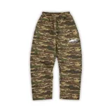 Pantalón de jogging Corteiz con estampado de camuflaje digital en tonos verdes y marrones. Presenta cintura elástica y un logo gráfico ovalado estampado en color blanco en la pierna izquierda.