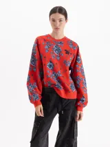 Buzo de algodón con cuello redondo y mangas largas, estampado con diseño de flores y alfileres de gancho sobre fondo rojo. Presenta puños y ruedo en tejido elástico al tono.