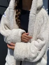 Abrigo tipo cárdigan de peluche en color crudo, con diseño holgado, capucha amplia y mangas largas con puños versátiles. Presenta una textura suave y mullida, ideal para un estilo casual y confortable.