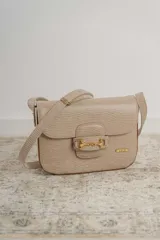 Cartera tipo bandolera de cuero con textura grabada, color beige. Presenta un detalle de herraje metálico dorado en la solapa frontal y placa con logo de la marca. Incluye correa ajustable para llevar al hombro o cruzada.