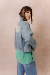 Campera de denim con lavado claro y efecto desgastado. Presenta cuello camisero, cierre frontal con botones metálicos, dos bolsillos frontales con tapa y botón, y puños abotonados.