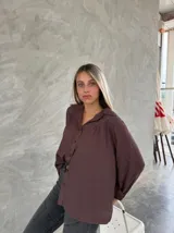 Camisa de corte holgado color marrón, con cuello clásico, cierre frontal de botones y detalle de bordado en la zona de los hombros y canesú. Presenta mangas largas con volumen.