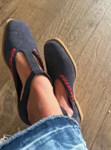 Ballerinas de gamuza color marrón oscuro con diseño tipo T-strap y detalles de costuras decorativas en color verde oliva. Cuentan con cierre de pulsera al tobillo y suela plana.