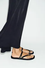 Sandalia plana de piel negra, con diseño de tiras que separan el dedo y detalle de hebilla metálica dorada en el empeine.