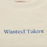 Remera de algodón color hueso con corte regular. Presenta un pequeño estampado con el texto 'Wasted Talent' en el centro del pecho y un detalle de costura en forma de 'W' en la manga izquierda.
