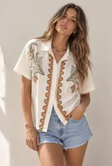 Camisa beige de manga corta con cuello y cierre frontal de botones. Presenta delicados bordados de hojas y flores en tonos verdes y naranjas a los lados, y una guarda ornamental ondulada alrededor de la botonera.